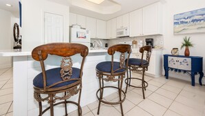 A kitchen with white cabinets, a white countertop, and a blue cabinet with floral designs. There are two ornate wooden chairs with blue cushions. - Lighthouse 215 3 Bedroom Condo (Gulf Shores)