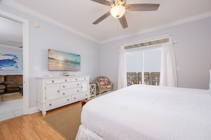 Une chambre à coucher comprenant un lit, un ventilateur de plafond, une commode, une chaise, un tableau et une fenêtre avec des rideaux.