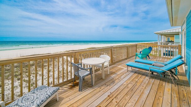 A wooden deck with a table and chairs, a lounge chair, and a view of the beach. - Funspot 1713 5 Bedroom Home (Gulf Shores)