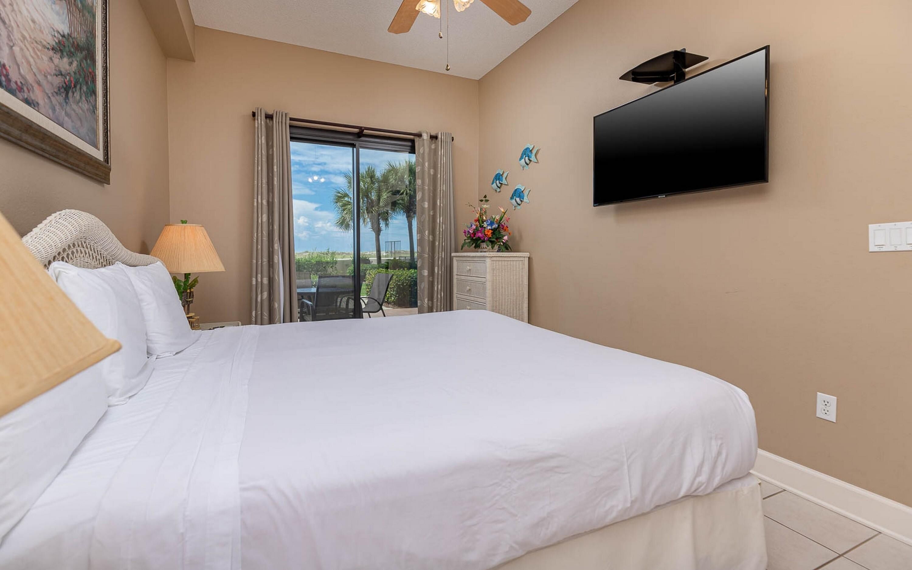 A hotel room with a large bed, a flat-screen TV mounted on the wall, a window with curtains, a small table with a lamp, and a decorative wall piece.