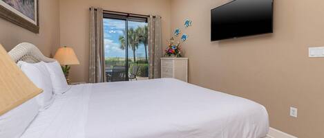 A hotel room with a large bed, a flat-screen TV mounted on the wall, a window with curtains, a small table with a lamp, and a decorative wall piece.