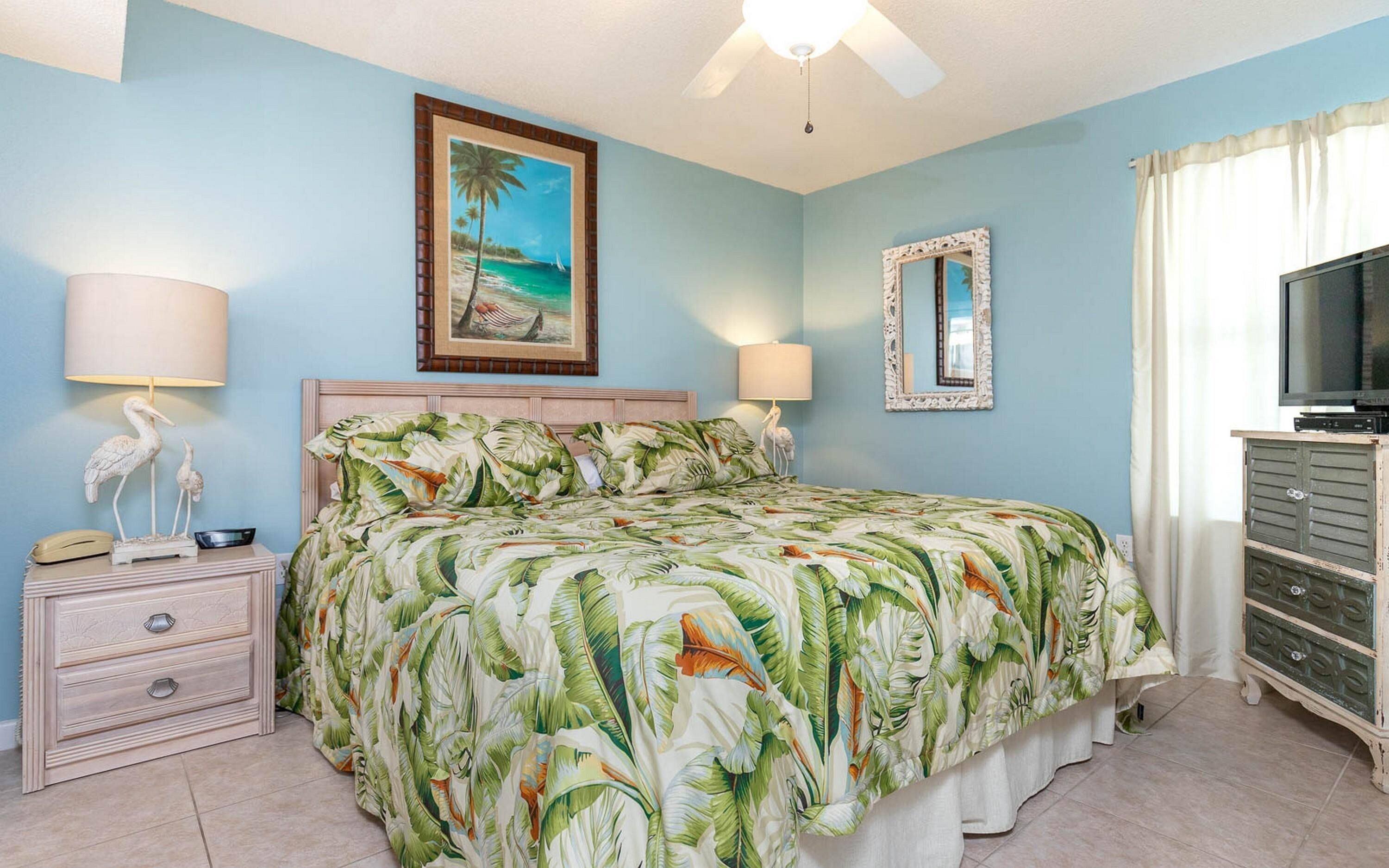 A bedroom with a bed, two bedside lamps, a painting, a mirror, a television, and a dresser.