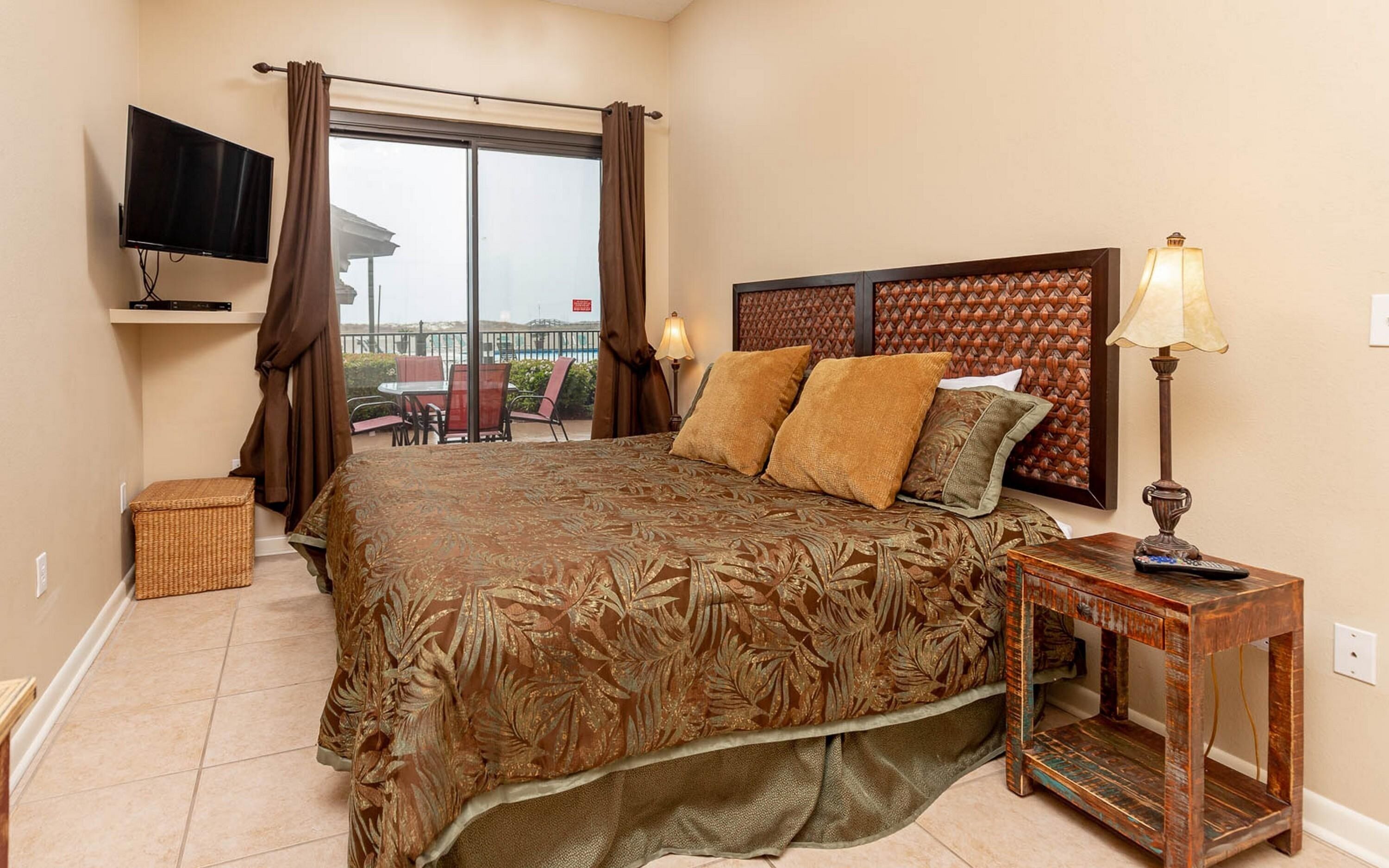 A bedroom with a large bed, a television mounted on the wall, a window with curtains, a nightstand with a lamp, and a view of the outdoors.