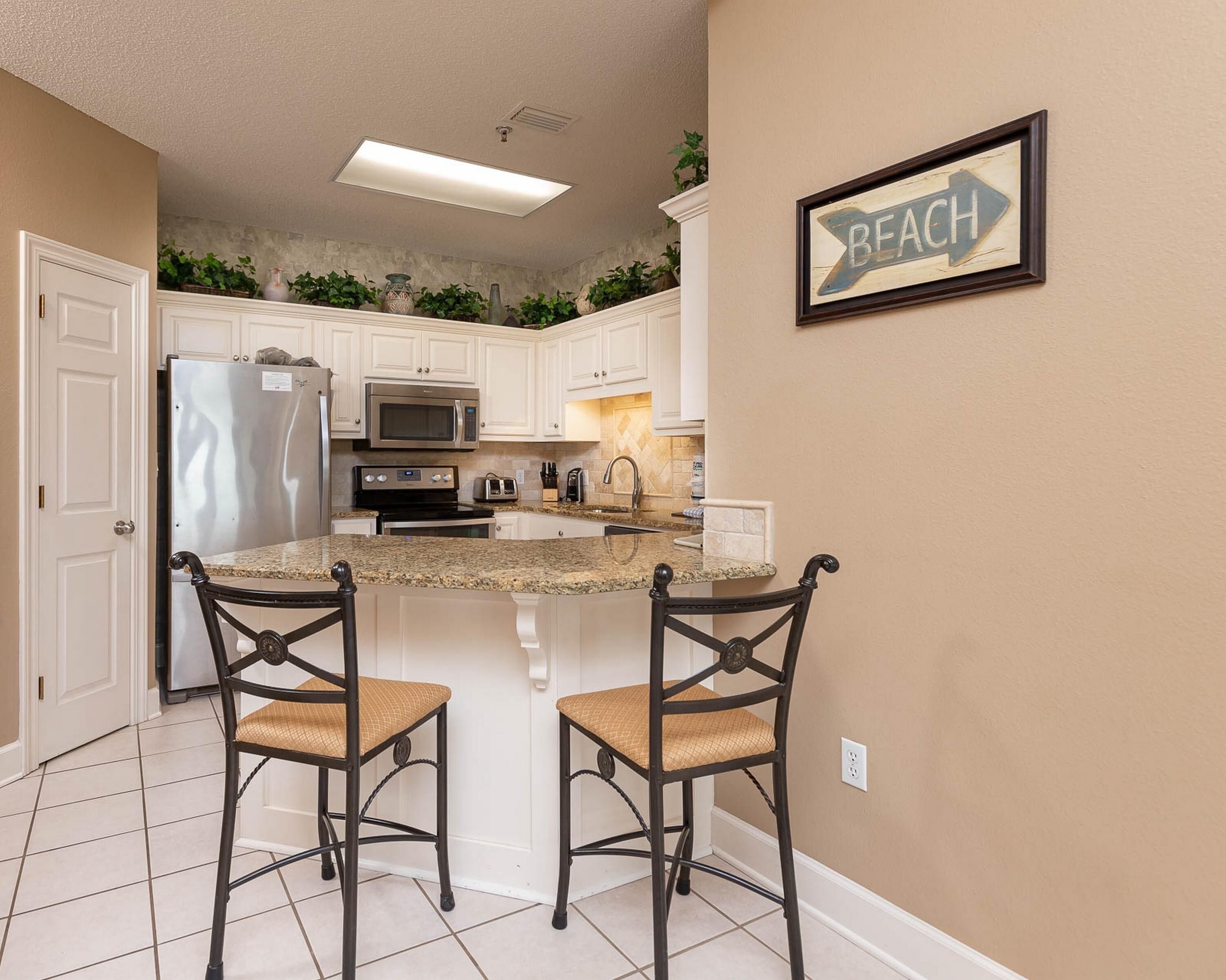 A kitchen with white cabinets, a stainless steel refrigerator, a microwave, and a double sink. There are two bar stools with woven seats.