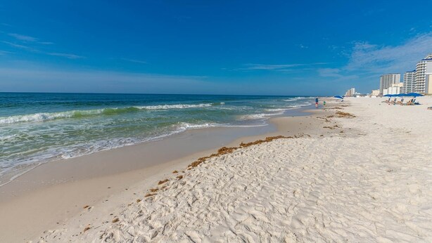 A beach with white sand, ocean waves, and a clear blue sky. There are buildings and umbrellas in the background.