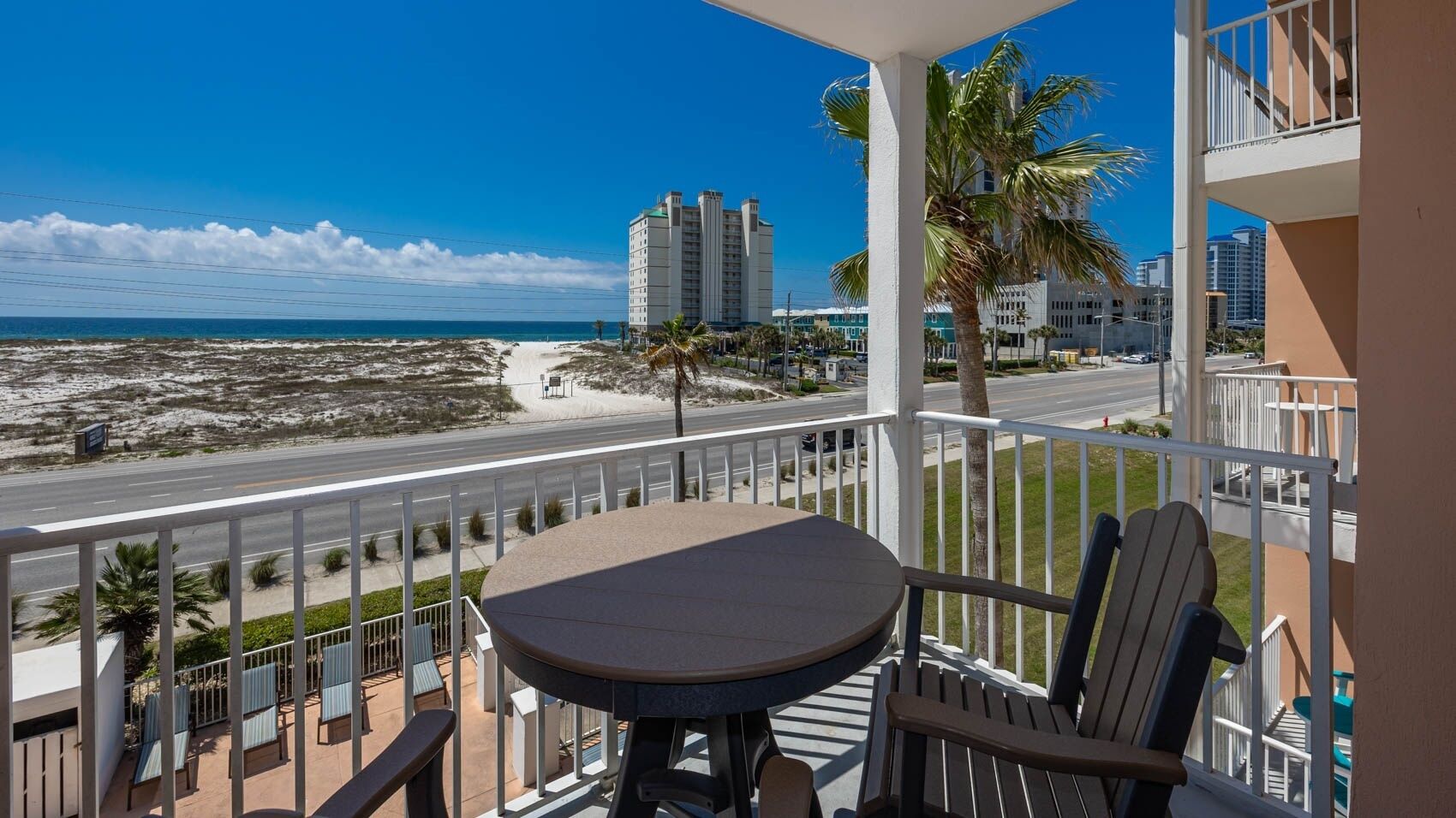 Een balkon met een tafel en stoelen, uitkijkend op een strand en een weg met gebouwen op de achtergrond.
