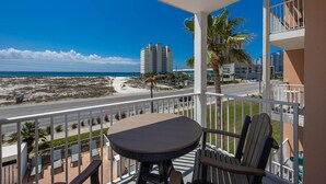 A balcony with a table and chairs, overlooking a beach and a road with buildings in the background.