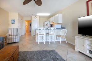 A modern living room with a TV, a sofa, a coffee table, and a dining area with chairs and a kitchen in the background. - Grand Beach Resort 308 1 Bedroom Condo (Gulf Shores)