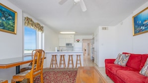 A living room with a red sofa, wooden coffee table, and a dining area with a wooden table and chairs. A kitchen is visible in the background. - Royal Palms 708 2 Bedroom Condo (Gulf Shores)