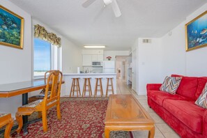A living room with a red sofa, wooden coffee table, and a dining area with a wooden table and chairs. A kitchen is visible in the background. - Royal Palms 708 2 Bedroom Condo (Gulf Shores)
