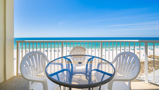 A balcony with a glass table and white chairs, overlooking a beach with a clear blue sky.