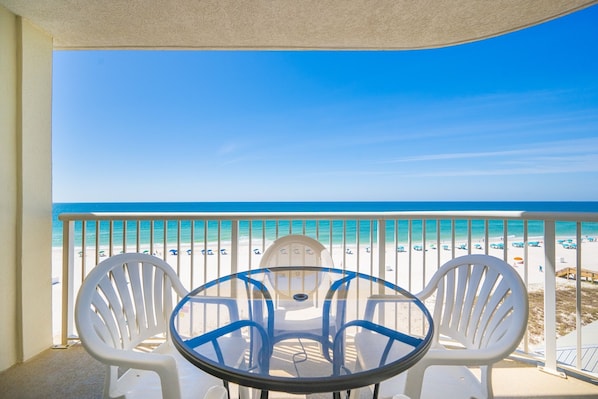 A balcony with a glass table and white chairs, overlooking a beach with a clear blue sky.
