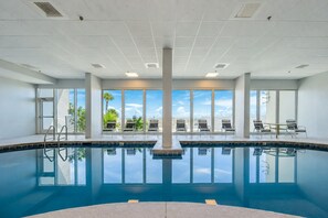 A large indoor swimming pool with clear blue water, surrounded by lounge chairs and a view of palm trees and a clear sky.