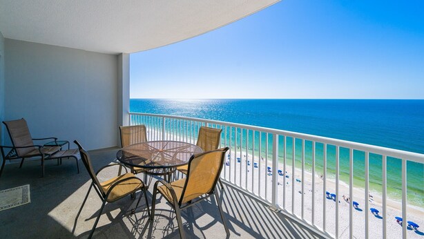 A balcony with a table and chairs, a view of the ocean, and a beach with umbrellas.
