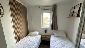 Una stanza con due letti singoli, una finestra con tende, un piccolo comodino in legno e una mensola a muro con delle foto.