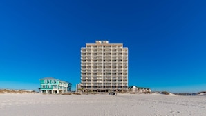 A tall hotel with multiple balconies, a beach in the foreground, and a clear blue sky. - Royal Palms 1207 1 Bedroom Condo (Gulf Shores)