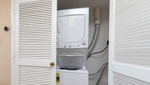 A compact laundry room with a front-loading washer and dryer, a door with a handle, and a visible hose. - Royal Palms 1207 1 Bedroom Condo (Gulf Shores)