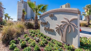 A sign for Royal Palms with a palm tree design, surrounded by a landscaped garden with various plants and flowers. - Royal Palms 1207 1 Bedroom Condo (Gulf Shores)