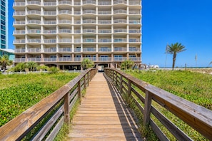A wooden boardwalk leading to a multi-story hotel with balconies, surrounded by greenery and a clear sky.