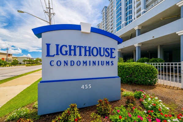 Un letrero para Lighthouse Condominiums que muestra claramente la entrada del edificio y un área ajardinada con flores.
