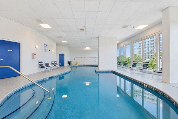 A large indoor swimming pool with clear blue water, surrounded by lounge chairs and a view of a high-rise building.