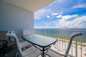 A balcony with a glass table and chairs, overlooking a beach with umbrellas and people.