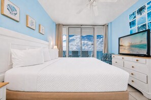 Une chambre d’hôtel avec un grand lit, une télévision, un balcon offrant une vue sur le ciel et les nuages, et des œuvres d’art accrochées aux murs.
