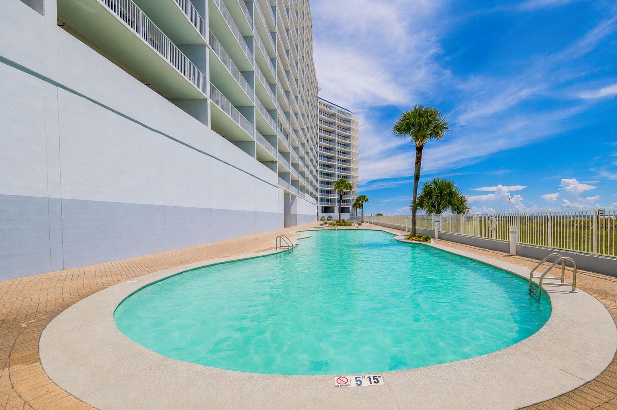 A swimming pool with a clear blue water, surrounded by a white fence, and a building with balconies in the background.