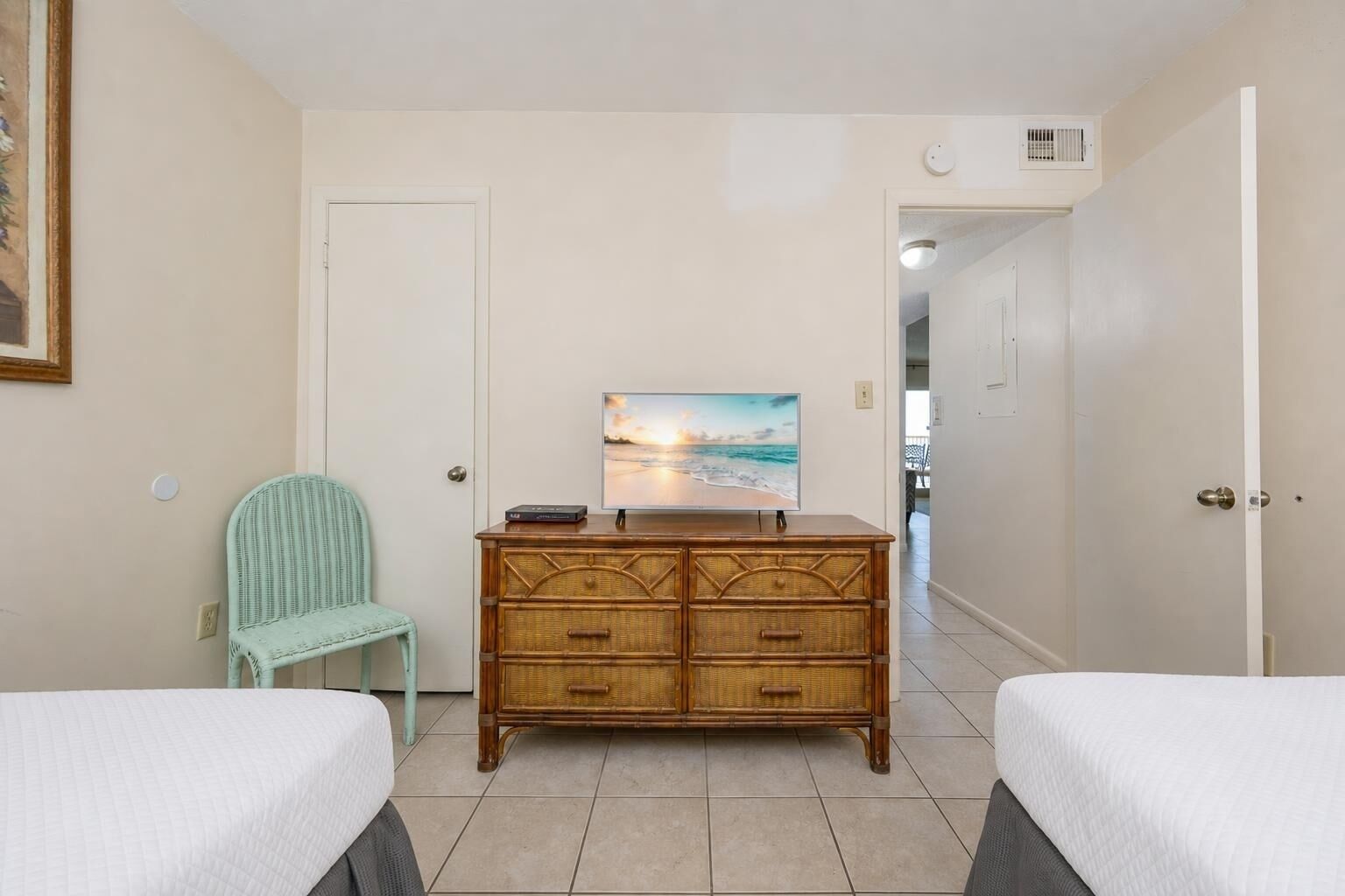 A hotel room with two beds, a wicker chair, a wooden dresser, and a painting of a beach scene.