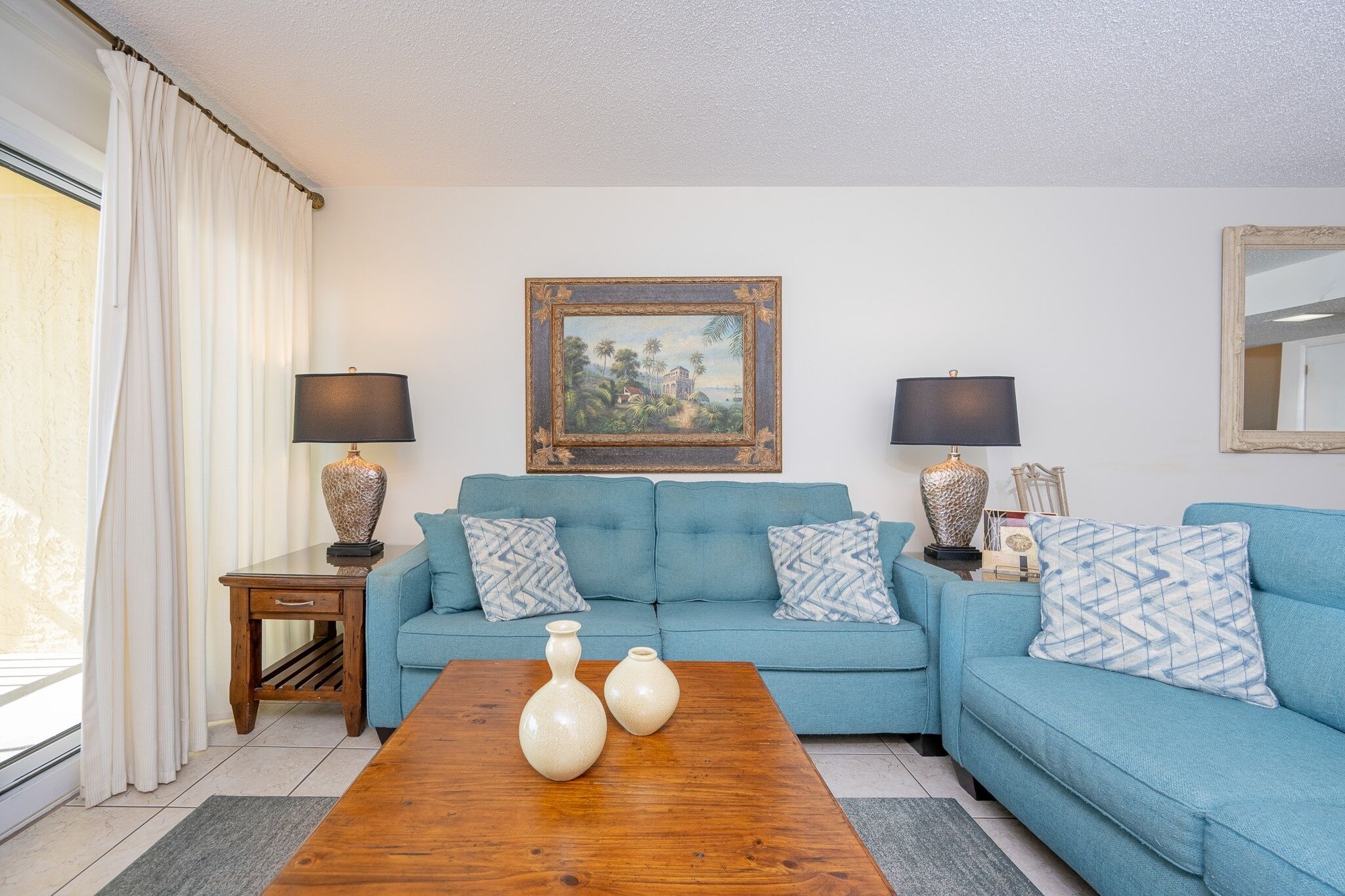 A living room with a blue sofa, two side tables with lamps, a wooden coffee table with vases, a painting on the wall, and a window with curtains.