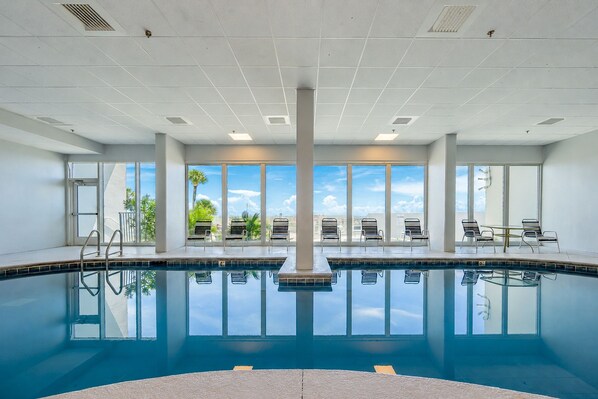 A large indoor swimming pool with clear blue water, surrounded by lounge chairs and a view of palm trees and a clear sky.