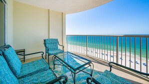 A balcony with a glass table and blue patterned furniture, overlooking a beach with a clear view of the ocean.