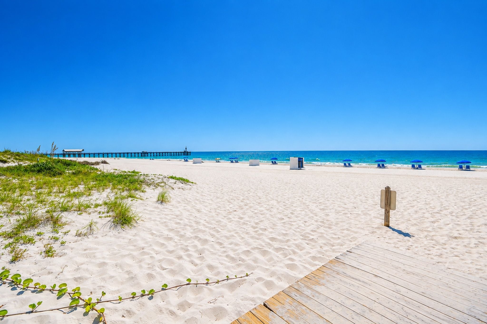 Een strand met een houten looppad dat naar het water leidt, heldere luchten en een pier in de verte.