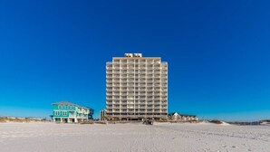 A tall hotel with multiple balconies, a beach in the foreground, and a clear blue sky.