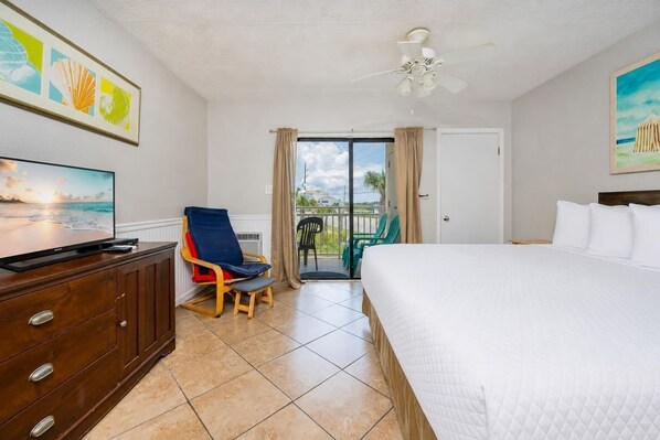 A hotel room with a bed, a TV, a chair, a ceiling fan, and a balcony with chairs. - Sugar Beach 177 1 Bedroom Condo (Gulf Shores)