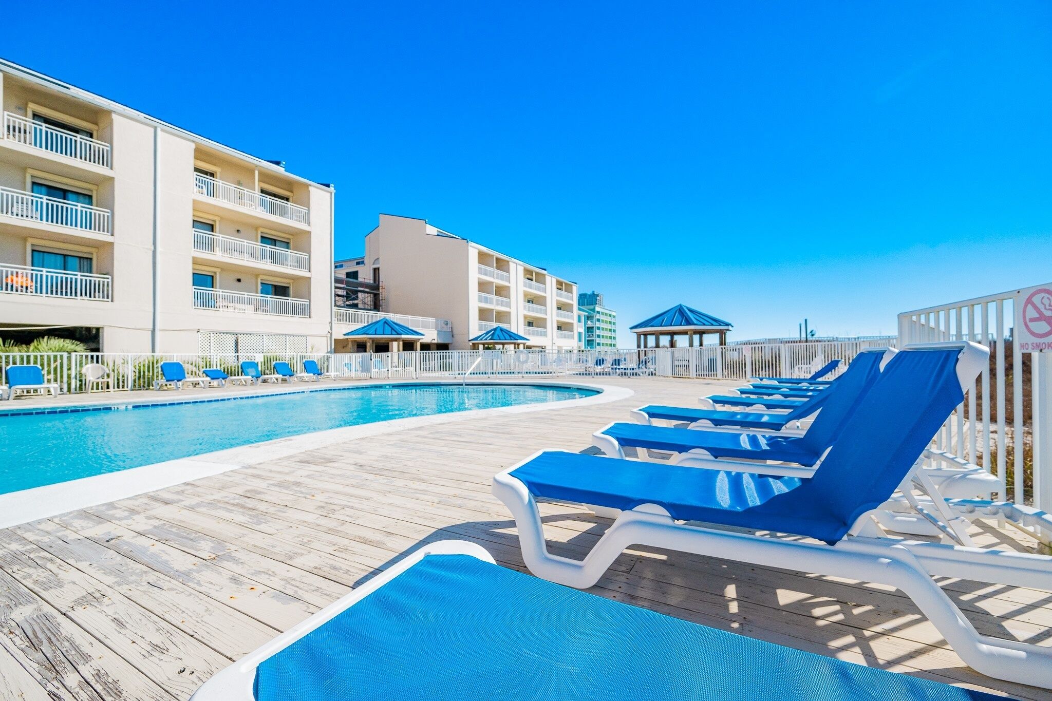 Zona junto a la piscina con sillas reclinables azules, terraza de madera y un edificio con varios balcones.