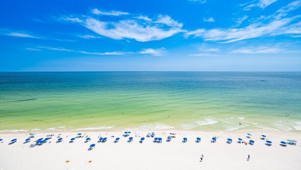 A beach with white sand, clear blue water, and a few umbrellas. - Crystal Shores West 807 2 Bedroom Condo (Gulf Shores)