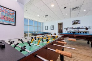 A room with two foosball tables, a large window with a view, and a wall with a framed picture. - Crystal Shores West 807 2 Bedroom Condo (Gulf Shores)