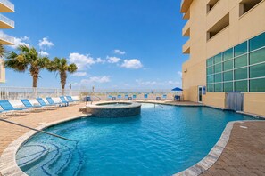 A swimming pool with lounge chairs and a hot tub, surrounded by palm trees and a building with large windows. - Crystal Shores West 807 2 Bedroom Condo (Gulf Shores)