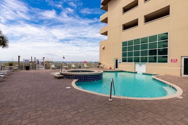 A pool area with a hot tub, lounge chairs, and a building with large windows. - Crystal Shores West 807 2 Bedroom Condo (Gulf Shores)