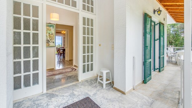 A covered outdoor area with a wooden ceiling, a white wall with green shutters, and a tiled floor.
