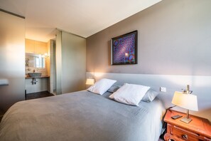 Une chambre d’hôtel avec un lit, des lampes de chevet, une œuvre d’art accrochée au mur et une salle de bain visible à travers une porte ouverte.