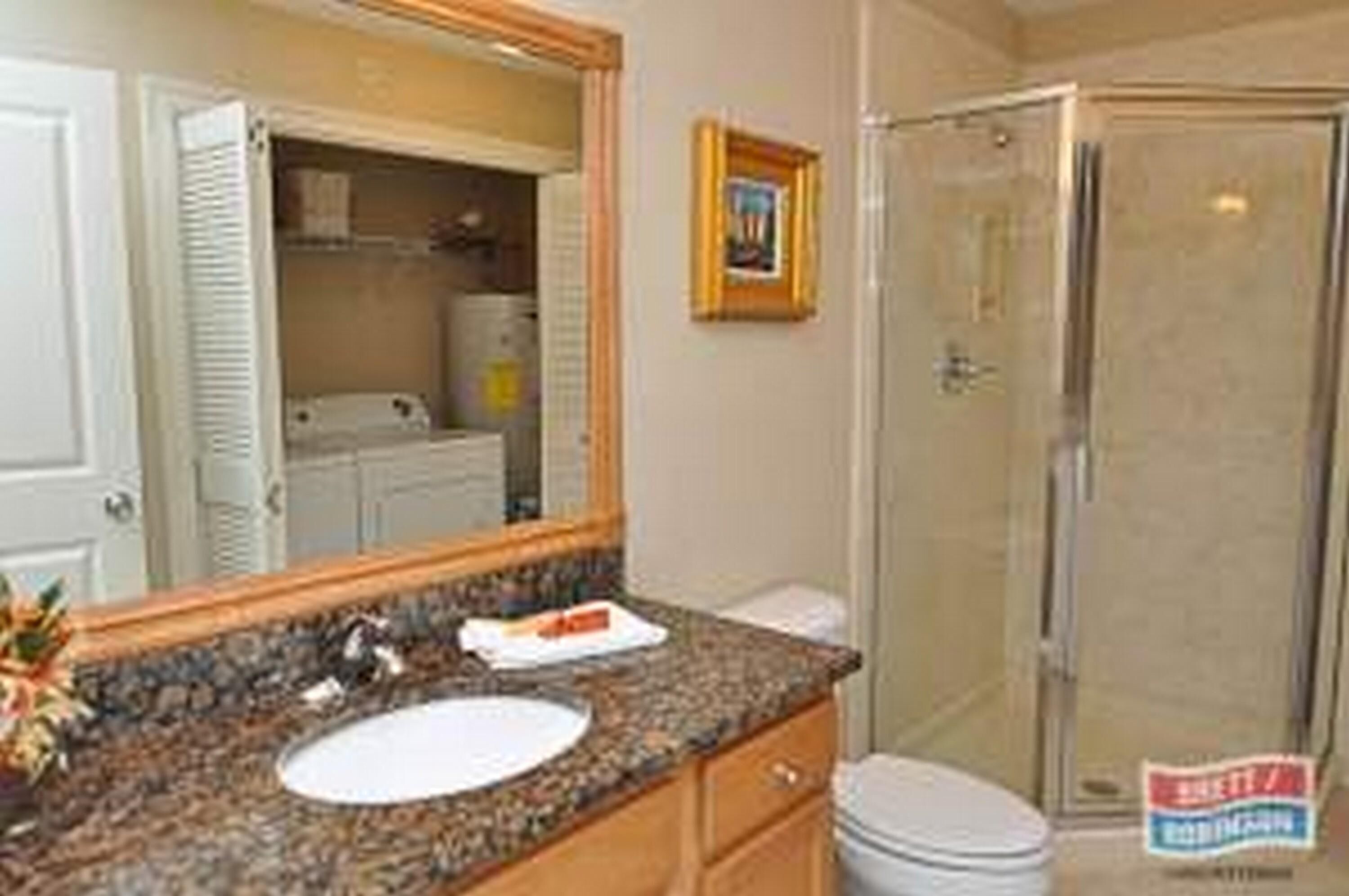 A bathroom with a granite countertop sink, wooden cabinets, a framed picture on the wall, and a glass shower enclosure.