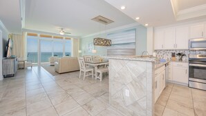 A modern kitchen with a central island, a dining area with a view, and a living room with a sofa and a TV.