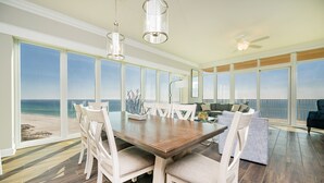 A spacious dining area with a large wooden table and white chairs, overlooking a beach.