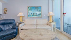 A living room with a blue leather sofa, a beige sofa, a wooden coffee table, and two lamps on side tables. A painting of a beach scene hangs on the wall. - Lighthouse 403 1 Bedroom Condo (Gulf Shores)