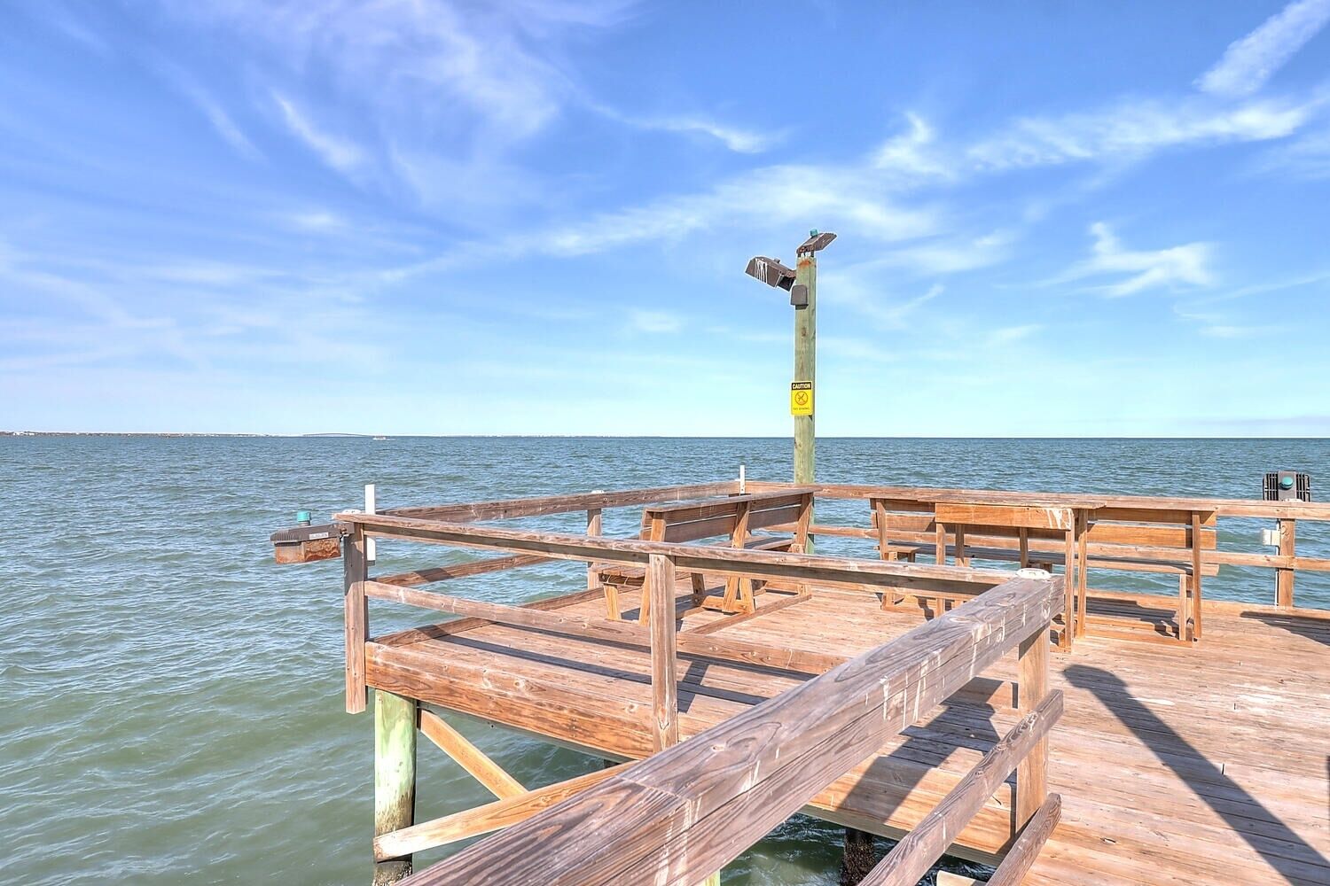 A wooden pier extending into calm waters with a caution sign, benches, and a clear sky.