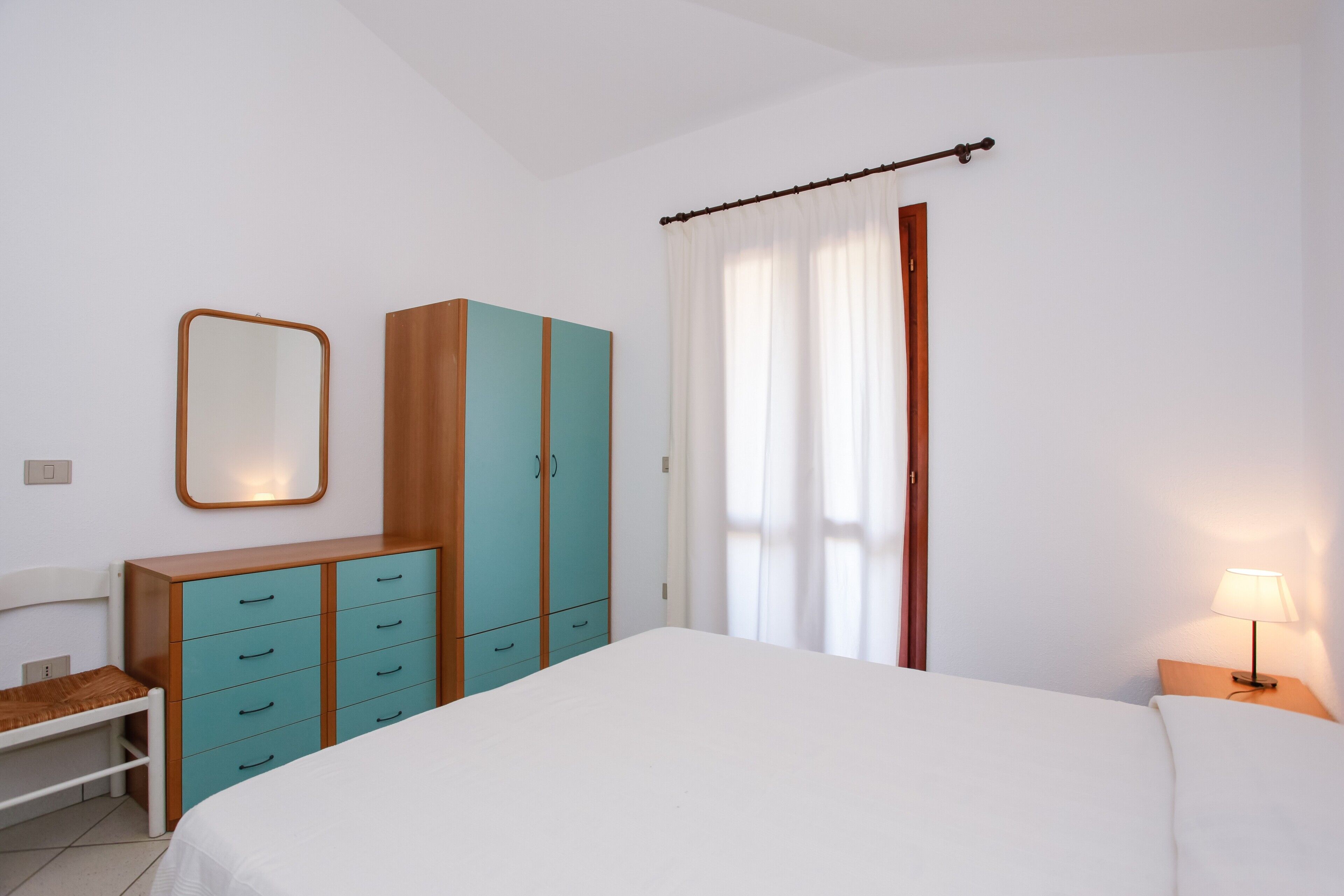 A bedroom with a bed, a wooden wardrobe, a mirror, a nightstand with a lamp, and a window with curtains.