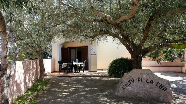 Una piedra con la inscripción 'COLTU DI LA ENA', una mesa y sillas bajo un árbol, y la entrada de un edificio.