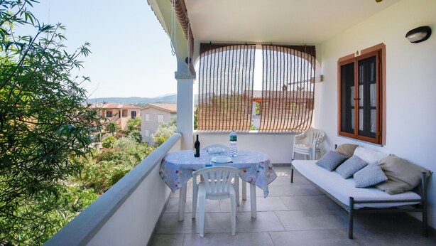 A balcony with a table, chairs, a sofa, and a window with a view of buildings and greenery. - Vacation Home "Trilo Mare" with Sea View, Private Terrace & Wi-Fi (Budoni)